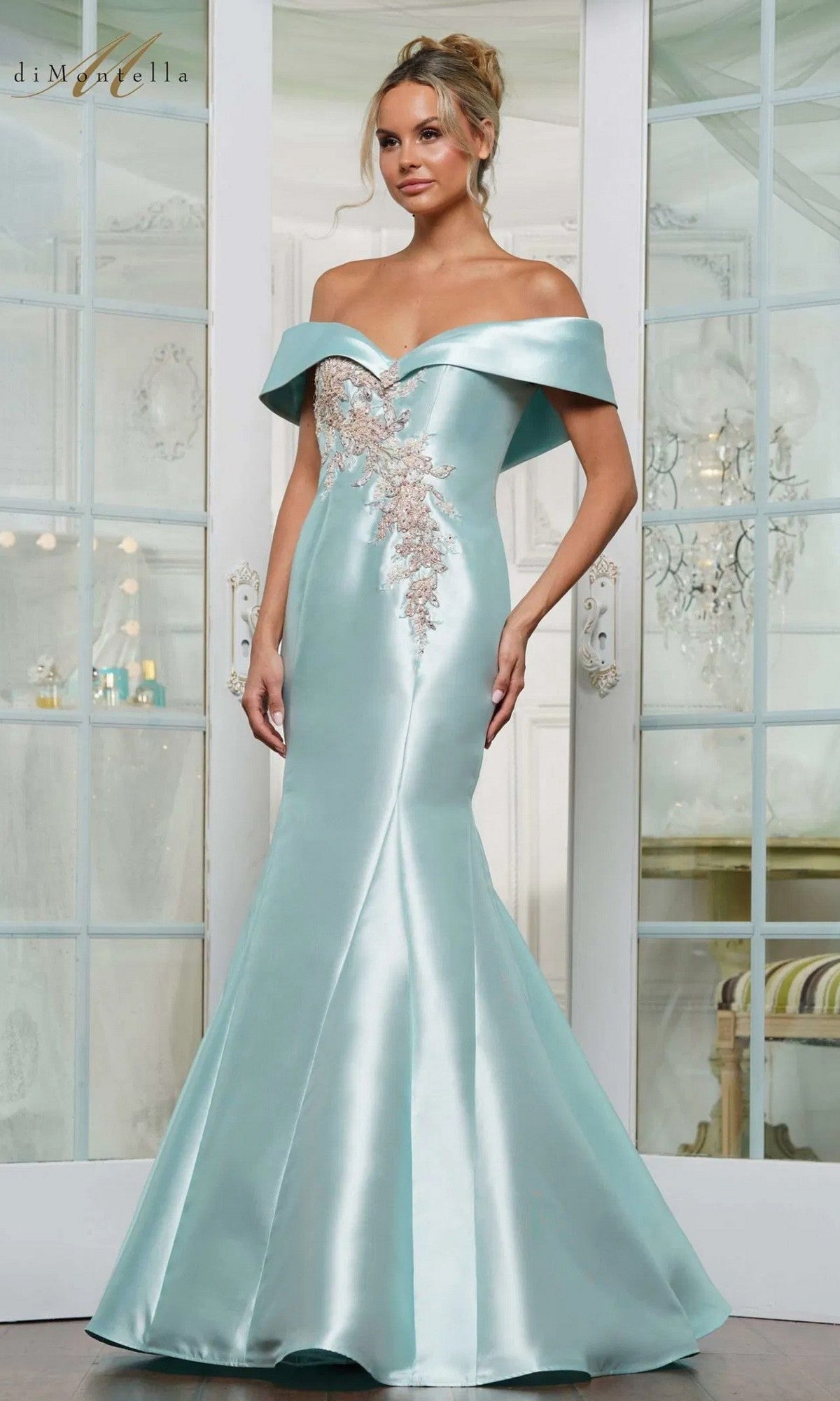 A woman stands indoors in front of glass doors, wearing the Rina Di Montella RD3257—a long light blue, off-the-shoulder formal gown featuring beaded floral embroidery and lace appliqué.